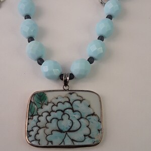 May include: A light blue beaded necklace with a silver clasp and a rectangular pendant. The pendant features a floral design in shades of blue and green, set against a white background. The necklace is a delicate piece of jewelry.