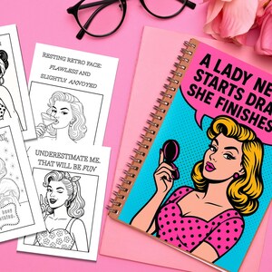 May include: A spiral-bound notebook with a pop art illustration of a woman holding a compact mirror, with the text "A LADY NEVER STARTS DRAMA - SHE FINISHES IT." Also, there are several black and white retro-style cards.