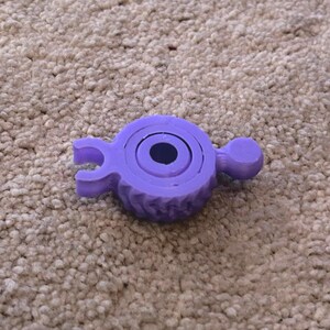 Può includere: Un giocattolo di plastica viola con un anello al centro e due braccia su entrambi i lati. Il giocattolo è progettato per essere un giocattolo fidget.