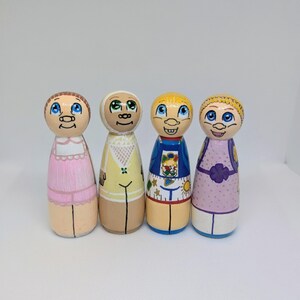 May include: Four hand-painted wooden peg dolls. Each doll features a unique design, including painted faces, clothing, and hairstyles. The dolls are in various colours, including pink, yellow, blue, and purple. The dolls are standing upright.
