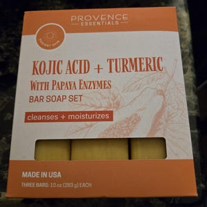 Puede incluir: Una caja de tres barras de jabón de ácido kójico y cúrcuma con enzimas de papaya. La caja es naranja y blanca con un gráfico de sol y el texto "Piel radiante". Las barras de jabón son amarillas y son visibles a través de una ventana en la caja. La caja también dice "Limpia + Hidrata" y "Hecho en EE. UU.".