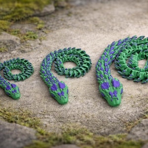 May include: Three articulated snake toys in varying sizes. The snakes are green with blue and purple accents. They are coiled on a textured, light brown surface with patches of green moss and small rocks.
