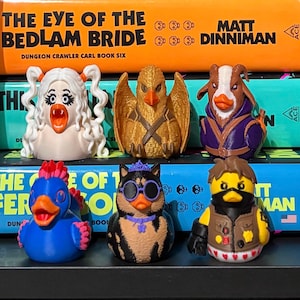 May include: Six small, stylized rubber duck figures arranged in two rows. The top row features a white-haired duck, a gold-winged duck, and a brown goat-like duck. The bottom row includes a blue duck, a black cat-like duck, and a tactical-themed duck.