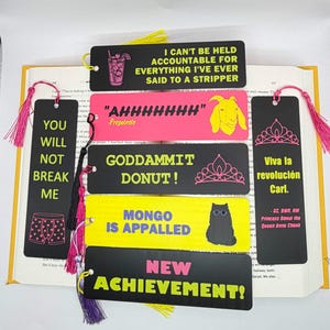 May include: Six rectangular bookmarks with tassels, featuring various colours and text. One black bookmark says I CAN'T BE HELD ACCOUNTABLE FOR EVERYTHING I'VE EVER SAID TO A STRIPPER. Another pink one says AHHHHHHHH with a goat illustration. A black one says GODDAMMIT DONUT! with a tiara. A yellow one says MONGO IS APPALLED with a cat. A black one says YOU WILL NOT BREAK ME with underwear. A black one says Viva la revolución Carl. with a tiara and NEW ACHIEVEMENT!.
