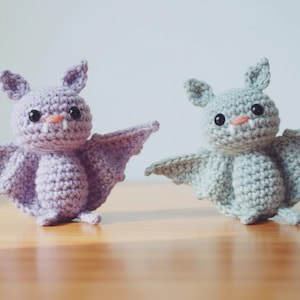 May include: Two handmade crochet bats in shades of purple and grey. Each bat has large black eyes, small orange noses, and tiny fangs. The bats have outstretched wings and are displayed on a wooden surface.