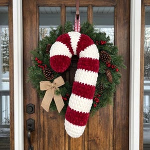 May include: A Christmas wreath with a large, plush candy cane decoration. The candy cane is red and white striped, and the wreath includes pine needles, pine cones, red berries, and a burlap bow. The wreath hangs on a wooden door.