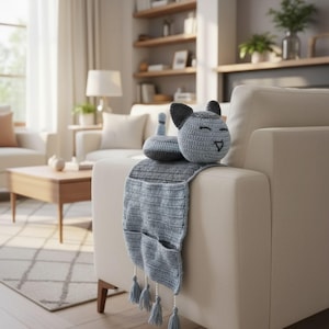 Op de afbeelding: Een gehaakte katvormige armleuning organizer in grijstinten en blauw. De kat heeft een lachend gezicht en oren. De organizer heeft zakken en kwastjes, ontworpen om over een bankarm te draperen.