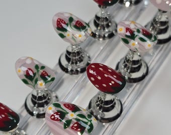 May include: A collection of nail art samples featuring various designs. Some nails are painted with a red mushroom design with white dots, while others showcase strawberries and white flowers on a pink background. The nails are displayed on clear stands.
