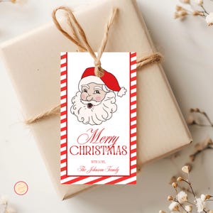 May include: A rectangular Christmas gift tag with a Santa Claus illustration and red and white striped borders. The tag reads "Merry Christmas" and includes a space for a family name. The tag is tied to a wrapped gift with twine.