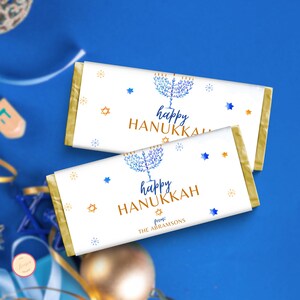 May include: Two chocolate bar wrappers with gold edges and white backgrounds. The wrappers feature blue and gold illustrations of a menorah, stars, and the words "happy HANUKKAH". The bottom wrapper includes the text "from: THE ABRAMSONS".