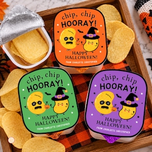 May include: Three snack containers with orange, green, and purple lids featuring cartoon potato chips, the text chip, chip HOORAY! HAPPY HALLOWEEN! FROM CHARLOTTE CHRISTOPHER, and scattered potato chips on a wooden tray with a plaid fabric accent.