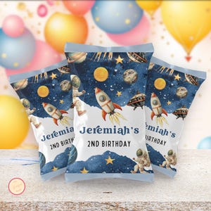 Puede incluir: Tres bolsas de aperitivos con un diseño espacial. Las bolsas presentan cohetes, planetas, astronautas y el texto "Jeremiah's 2nd Birthday" sobre un fondo azul oscuro estrellado. Las bolsas están selladas en la parte superior y tienen una base blanca.
