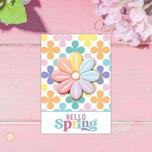 Könnte beinhalten: Ein pastellfarbener blütenförmiger Keks auf einer Karte mit floralem Muster. Auf der Karte steht in bunten Buchstaben "Hello Spring". Der Hintergrund ist eine rosa Holzoberfläche.
