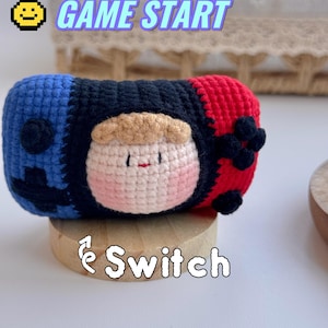 May include: Crocheted Nintendo Switch case in blue, black, and red, featuring a cartoon character face. The case is on a wooden stand, with the words "GAME START" and "Switch" in the background.