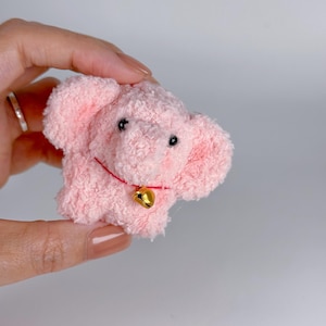 May include: A small, fuzzy, light pink plush elephant with large ears and a textured, yarn-like surface. The elephant features two small black bead eyes and a thin red cord around its neck, which holds a single, shiny, gold-colored metal bell.