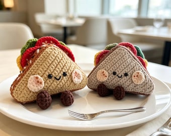 May include: Two triangular crochet sandwich plushies with smiling faces, black bead eyes, and pink cheeks. Each features layers of green, red, and yellow yarn representing fillings, with small brown crocheted feet attached to the bottom edges.