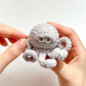 以下が含まれることがあります： 柔らかい質感の糸で作られた、淡いグレーの小さなタコのぬいぐるみ。丸みを帯びた頭部、8本のカールした触手、そして折りたたまれた眉の下に配置された、黒い瞳を持つ2つの小さな白い楕円形の目が特徴です。