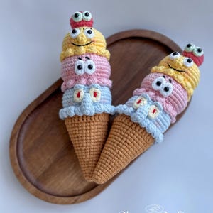 May include: Two handmade crochet ice cream cones with smiling faces and googly eyes. Each cone features three scoops in yellow, pink, and blue, atop a brown cone. The cones are placed on a wooden tray. The text "Cheney Crafts" is visible.