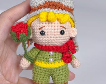 May include: A small crochet doll featuring a character with yellow hair, wearing a brown and beige aviator-style hat, a red scarf, and a green outfit with a yellow waistband. The doll has two white pearl-like buttons and holds a red rose on a green stem.