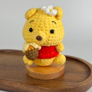May include: A yellow crocheted bear figure wearing a red shirt, featuring a white flower on its head and holding a brown textured honey pot. The bear has black eyes, a dark brown nose, and is mounted on a small, round, light-coloured wooden base.