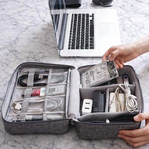 Könnte beinhalten: Ein grauer, mit Reißverschluss versehener Elektronik-Organizer mit mehreren Fächern, der verschiedene Kabel, Ladegeräte und eine Festplatte enthält. Ein Laptop, eine Maus und eine Brieftasche befinden sich im Hintergrund. Der Organizer wird geöffnet gehalten.