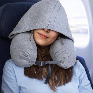 May include: A gray travel pillow with a hood is worn by a person on an airplane. The pillow wraps around the neck and has a hood that covers the head and face. The person is wearing a light blue shirt.