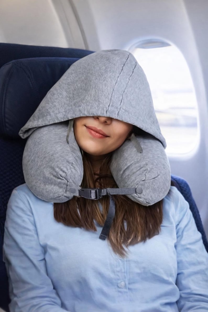 May include: A gray travel pillow with a hood is worn by a person on an airplane. The pillow wraps around the neck and has a hood that covers the head and face. The person is wearing a light blue shirt.