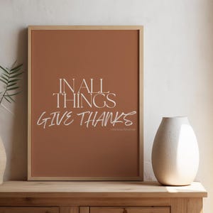 Op de afbeelding: Een ingelijste print met de tekst "IN ALL THINGS GIVE THANKS" in wit schrift tegen een terracotta achtergrond. De print wordt weergegeven op een houten kast, naast een textuurvaas met groen en een witte keramische vaas. Een raam met uitzicht op de oceaan is op de achtergrond.