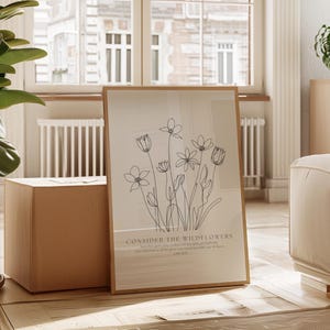 Può includere: Una stampa artistica incorniciata con un disegno a tratto di fiori selvatici in inchiostro nero. La stampa ha uno sfondo chiaro con il testo "Consider the wildflowers". La cornice è di colore marrone chiaro. L'opera d'arte è esposta in una stanza ben illuminata.