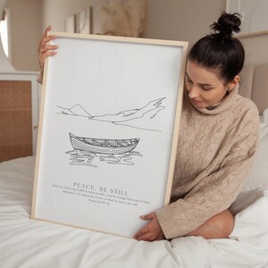 Pode incluir: Uma impressão artística emoldurada com um desenho minimalista de um barco na água e montanhas. A obra de arte em preto e branco inclui as palavras "PEACE, BE STILL" e um versículo bíblico. A moldura é de madeira clara.