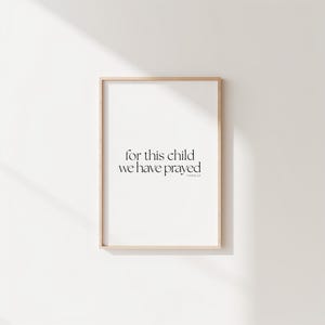 Può includere: Stampa incorniciata con la scritta "for this child we have prayed" in tipografia minimalista nera. Il testo è centrato su sfondo bianco, all'interno di una cornice in legno chiaro. La stampa è appesa a una parete bianca.