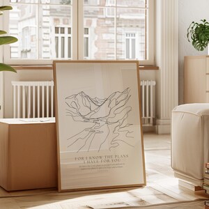 May include: A framed art print with a minimalist line drawing of mountains and a river. The artwork is in a light brown wooden frame and includes the text "FOR I KNOW THE PLANS I HAVE FOR YOU". The print is leaning against a brown cube.
