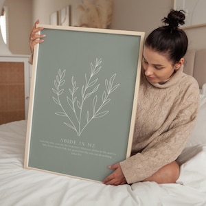 Könnte beinhalten: Gerahmter Kunstdruck mit salbeigrünem Hintergrund und weißer botanischer Zeichnung. Der Text "ABIDE IN ME" ist zu sehen, zusammen mit einem Zitat aus Johannes 15,5. Der Rahmen ist helles Holz, was eine minimalistische Ästhetik erzeugt. Für die Inneneinrichtung.
