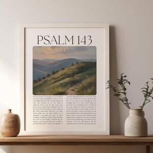 Könnte beinhalten: Gerahmter Kunstdruck mit dem Text "PSALM 143" über einem Landschaftsgemälde mit grünen Hügeln unter einem bewölkten Himmel. Der Druck befindet sich in einem weißen Rahmen und ruht auf einem Holztisch mit einer Vase und einem dekorativen Topf.
