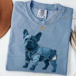 May include: A light blue Comfort Colors t-shirt featuring a graphic of a French Bulldog wearing a denim jacket. The shirt has a crew neck and short sleeves. A gold necklace with a pendant is draped across the shirt.