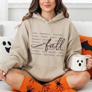 May include: A beige hooded sweatshirt featuring a dark brown graphic print of a list of fall-themed words including cozy sweaters, scarves, jeans, boots, apple cider, pumpkin patches, crunchy leaves, giving thanks, football, candy corn, and autumn.