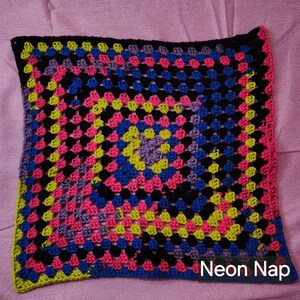 Könnte beinhalten: Ein quadratisches, handgefertigtes Häkelkissen mit einem lebendigen, geometrischen Muster. Das Design zeigt einen schwarzen Hintergrund mit Quadraten in Neonrosa, Gelb, Blau und Lila. Der Text "Neon Nap" ist unten rechts sichtbar.