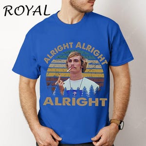 Puede incluir: Camiseta azul real con un gr&aacute;fico de un hombre y el texto "ALRIGHT ALRIGHT ALRIGHT" en un dise&ntilde;o circular. El dise&ntilde;o incluye una puesta de sol y &aacute;rboles. La palabra "ROYAL" est&aacute; impresa en la parte superior izquierda.