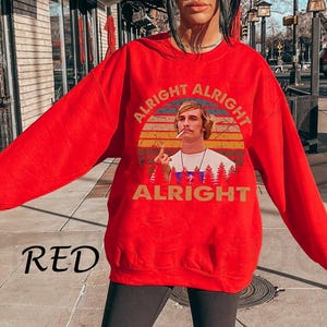 Puede incluir: Una sudadera roja con un gr&aacute;fico de un hombre fumando, sobre un dise&ntilde;o de puesta de sol. Las palabras "ALRIGHT" est&aacute;n impresas arriba y abajo de la imagen. La palabra "RED" est&aacute; impresa en la esquina inferior izquierda.