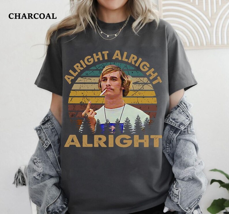 Puede incluir: Camiseta de color carb&oacute;n con la frase "ALRIGHT ALRIGHT ALRIGHT" y una imagen de un hombre con un cigarrillo. El dise&ntilde;o incluye un fondo estilo puesta de sol e im&aacute;genes de bosque.