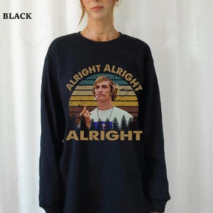 Puede incluir: Camiseta negra de manga larga con un gr&aacute;fico de un hombre fumando, el texto "ALRIGHT ALRIGHT ALRIGHT" en un dise&ntilde;o circular y una silueta de bosque. La camiseta es de cuello redondo.