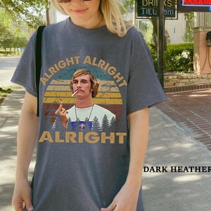 Puede incluir: Camiseta gris jaspeado oscuro con un dise&ntilde;o retro. El dise&ntilde;o presenta el texto "Alright Alright Alright" y una imagen de un hombre con un cigarrillo, sobre un fondo de puesta de sol y &aacute;rboles. Prenda de vestir informal.
