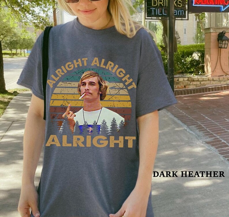 Puede incluir: Camiseta gris jaspeado oscuro con un dise&ntilde;o retro. El dise&ntilde;o presenta el texto "Alright Alright Alright" y una imagen de un hombre con un cigarrillo, sobre un fondo de puesta de sol y &aacute;rboles. Prenda de vestir informal.