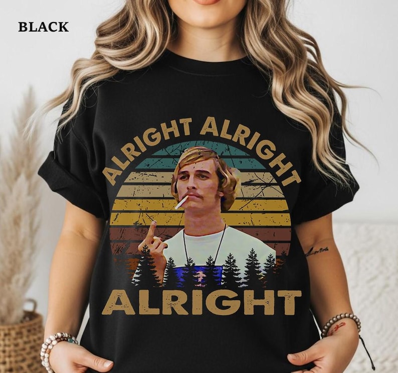 Puede incluir: Camiseta negra con un dise&ntilde;o retro. El dise&ntilde;o presenta las palabras "Alright Alright Alright" alrededor de un retrato de un hombre con un cigarrillo, sobre un fondo de puesta de sol y silueta de bosque.