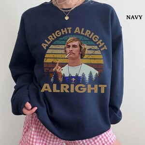 Puede incluir: Sudadera azul marino con un gr&aacute;fico retro de un hombre con un cigarrillo, sobre un fondo de puesta de sol. Las palabras "ALRIGHT ALRIGHT ALRIGHT" est&aacute;n arqueadas alrededor de la imagen. La palabra "NAVY" est&aacute; impresa en la esquina superior derecha.