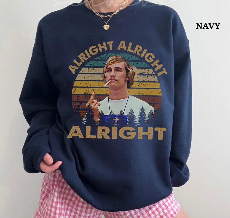 Puede incluir: Sudadera azul marino con un gr&aacute;fico retro de un hombre con un cigarrillo, sobre un fondo de puesta de sol. Las palabras "ALRIGHT ALRIGHT ALRIGHT" est&aacute;n arqueadas alrededor de la imagen. La palabra "NAVY" est&aacute; impresa en la esquina superior derecha.