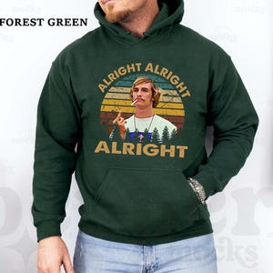 Puede incluir: Sudadera con capucha verde bosque con bolsillo delantero y un gr&aacute;fico de un hombre con las palabras "Alright Alright Alright" en un dise&ntilde;o retro. El texto "Forest Green" est&aacute; en la parte superior izquierda.