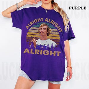 Puede incluir: Camiseta morada con un dise&ntilde;o gr&aacute;fico de un hombre y las palabras "Alright Alright Alright". El dise&ntilde;o incluye una puesta de sol y &aacute;rboles. La camiseta es de color morado liso.