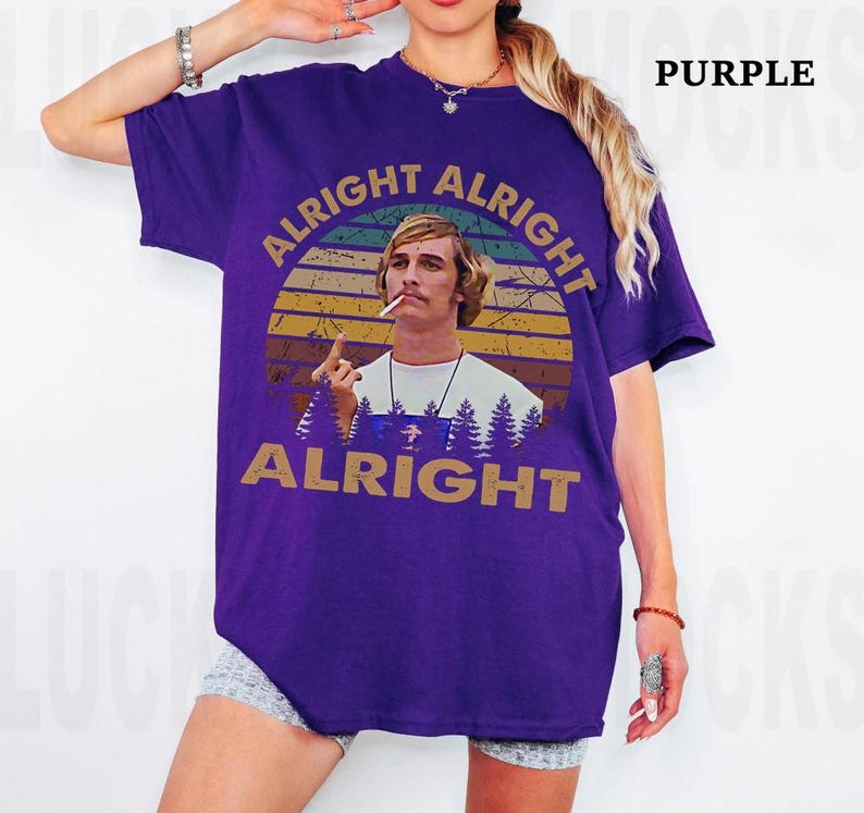 Puede incluir: Camiseta morada con un dise&ntilde;o gr&aacute;fico de un hombre y las palabras "Alright Alright Alright". El dise&ntilde;o incluye una puesta de sol y &aacute;rboles. La camiseta es de color morado liso.