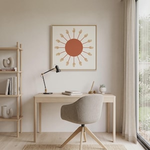 May include: A framed print depicting a sun design with a terracotta-coloured centre and beige hands radiating outwards. The artwork hangs above a light wood desk with a black desk lamp, books, and a beige swivel chair. A small plant is on the desk.